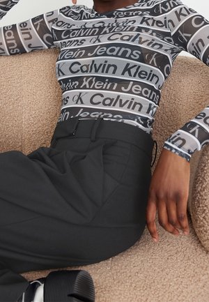 Person lehnt sich auf einem beigen, strukturierten Stuhl zurück und trägt ein schwarz-weißes, mit Calvin Klein Jeans gemustertes Netzoberteil und schwarze Hosen.