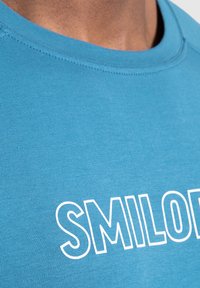 Tyrkysové tričko s kulatým výstřihem, na přední straně s výrazným bílým textem "SMILOD". Tkanina vypadá hladce, lehce a prodyšně.