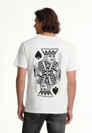 Mężczyzna w białej koszulce z dużą czarną grafiką króla pik na plecach, stojący na jednolitym tle.