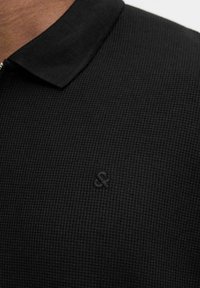 Sort polo-shirt med tekstureret strikstof, en klassisk krave og et præget logo på brystet, der understreger et rent design.