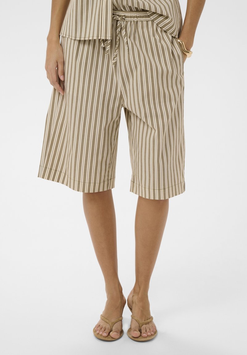 Femme portant un short beige et blanc à rayures verticales longueur genou avec une taille nouée, assorti à des sandales beiges en tongs et un bracelet en or.