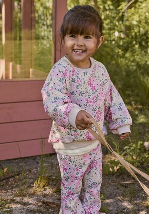 Naeratav väikelaps tüdruk, kes kannab roosat lillelist dressipluusi ja pükse, hoides beežikat rihma, seisab õues puidust ehitise lähedal roheluses.