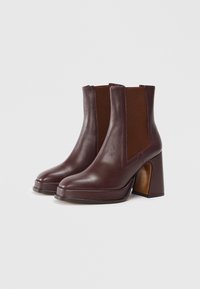 Bottes chelsea en cuir bordeaux avec panneaux latéraux élastiques et un talon haut épais marron. Texture lisse avec un design à bout pointu et des accents minimaux.