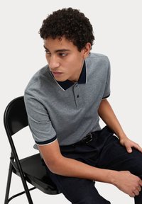 Grå polo skjorta med marinblå krage och ärmsnedd, med en treknäppnings placket. Modell sitter på en svart stol. Mörka byxor synliga.