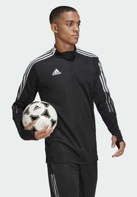 Schwarzes langärmeliges Sportoberteil mit weißem Adidas-Logo und drei weißen Streifen an den Ärmeln; Modell hält einen schwarz-weißen Fußball.