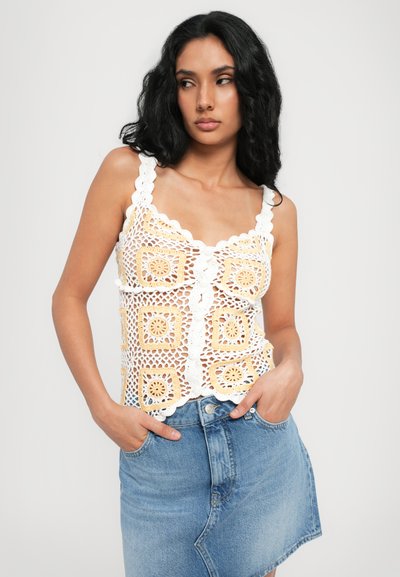Top croșetat cu modele pătrate albe și galbene, bretele umflate și nasturi în față. Asociat cu o fustă din denim albastru. Design texturat.