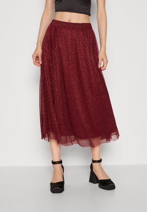 Kvinde iført en bourgogne glitrende midi-nederdel, sort cropped top og sorte ankelrem stiletter på ensfarvet baggrund.
