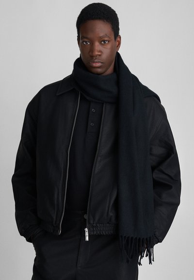 Joven vestido con una chaqueta de cuero negra, una camisa de botones negra, pantalones negros y una bufanda larga negra sobre un fondo gris.