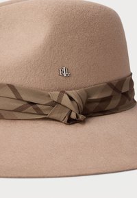 Beige színű filc fedora kalap barna kockás szövetpánttal, amelyet csomóra kötöttek, és elöl egy kis ezüst "RL" logó kitűző található.