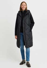 Črn prešiti dolg puffer brezrokavnik s kapuco, z zadrgo spredaj in stranskimi žepi, oblečen čez črno majico in modre kavbojke.