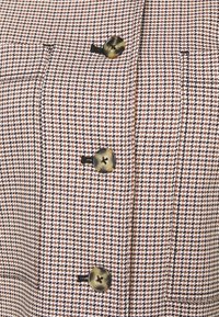 Gros plan sur une veste en tissu vichy marron et blanc avec trois grands boutons en écaille de tortue et deux poches passepoilées verticales.