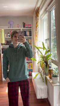 Πράσινη hoodie με κορδόνια και μπροστινή τσέπη, συνδυασμένη με καρό μπορντό παντελόνι. Στο φόντο υπάρχουν μια βιβλιοθήκη και εσωτερικά φυτά.