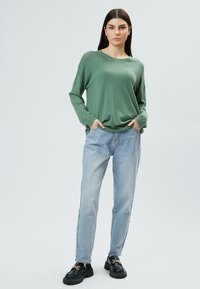Groene gebreide trui met een gestructureerd patroon op de schouders, gecombineerd met lichtblauwe rechte jeans en zwarte schoenen met een chunky zool.