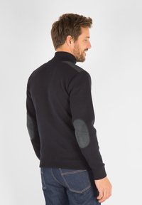 Pull en maille bleu marine avec un col montant, présenté avec des patches en daim noir aux coudes et des accents aux épaules. Poignets et ourlet côtelés.