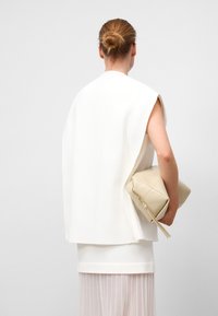 Top blanc sans manches au design fluide, porté par-dessus une robe blanche à couches, tenant un sac à main beige à motif géométrique.