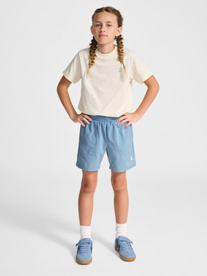 Młoda dziewczyna z warkoczami stoi z rękami na biodrach, ubrana w kremowy t-shirt, niebieskie szorty, białe skarpetki i niebieskie tenisówki na białym tle.