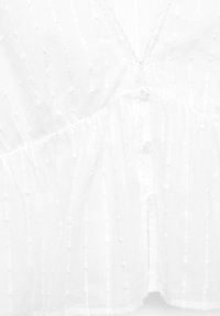 Blouse blanche avec un décolleté en V, tissu texturé avec des points en relief, des plis et des fermetures à boutons à l'avant. Matériau doux et léger.