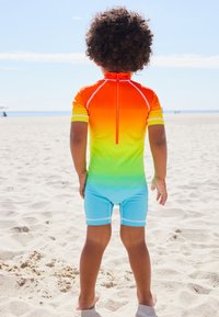 Brightly colored swim suit with an orange to green ombre design, short sleeves, a back zipper, and light blue shorts. Beach setting.