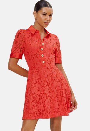Femme portant une robe courte rouge en dentelle avec des motifs floraux, des boutons dorés, un col et des poignets en satin, posant avec une main sur la hanche.