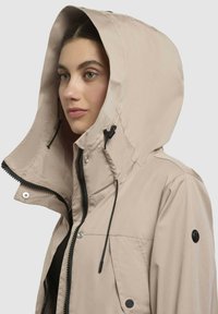 Giacca impermeabile beige con un ampio cappuccio, zip frontale, bottoni a pressione e coulisse. Presenta un accento nero sulla spalla.