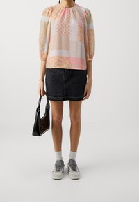 Femme portant un blouse en patchwork pastel, une jupe en denim noir, des baskets grises avec des chaussettes blanches, tenant un sac à main noir sur un fond uni.