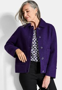 Giacca di lana viola con colletto, dotata di chiusura a bottoni e due tasche frontali. Indossata sopra una maglietta a motivo geometrico e pantaloni neri.