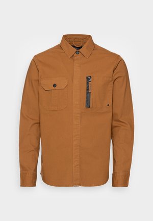 Chemise marron à manches longues avec col, deux poches poitrine - une avec rabat et bouton, l'autre avec fermeture éclair noire verticale.