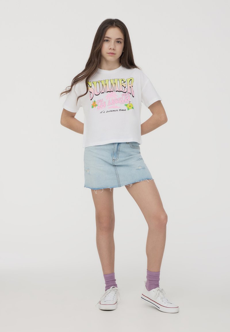 T-shirt bianco cropped con grafica colorata "SUMMER 1985 LOS ANGELES", abbinato a una mini gonna in denim chiaro e sneakers bianche con calzini viola.