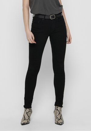 Person trägt schwarze Skinny-Jeans, schwarzen Gürtel, dunkelgraues Oberteil und knöchelhohe Stiefel mit Schlangenprint vor schlichtem hellem Hintergrund.