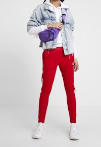 Jeansjacka med fransad nederkant, vit hoodie, röda sportbyxor med vita ränder och en lila axelväska. Vita sneakers fulländar outfiten.