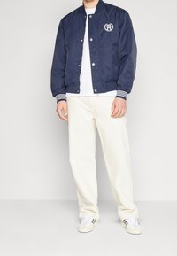 Navy bomber jacket with snap buttons, ribbed cuffs, and white logo. Paired with white pants and sneakers, showcasing a casual look.