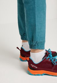Persoon in teal taps toelopende broek, witte sokken en maroon, oranje en blauwe Dachstein wandelschoenen staand op een lichte vloer.