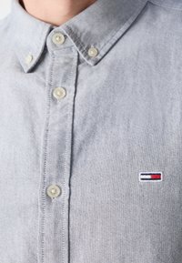 Detailní záběr na světle modrou košili s knoflíky, na levé straně hrudi je malá obdélníková nášivka v červené, bílé a modré barvě.