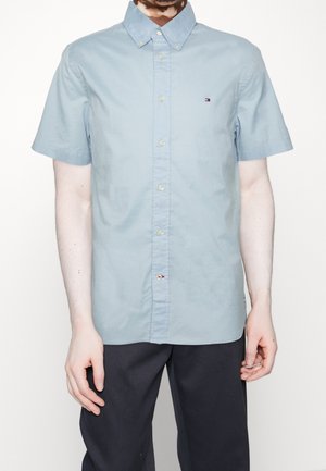 Mężczyzna ubrany w jasnoniebieską koszulę z krótkim rękawem na guziki i ciemne spodnie, stojący z ramionami swobodnie wzdłuż ciała na jednolitym tle.