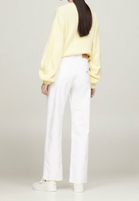 Suéter de punto amarillo con mangas abullonadas combinado con jeans anchos blancos. La modelo lleva zapatillas blancas; los detalles visibles incluyen un cinturón marrón y bolsillos.