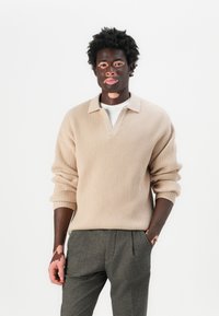 Maglione beige a coste con collo a V, indossato su una camicia bianca, abbinato a pantaloni grigi a trama.