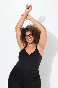 Woman with curly hair and black glasses wears black tank top and pants, smiling with arms raised and crossed above her head.
