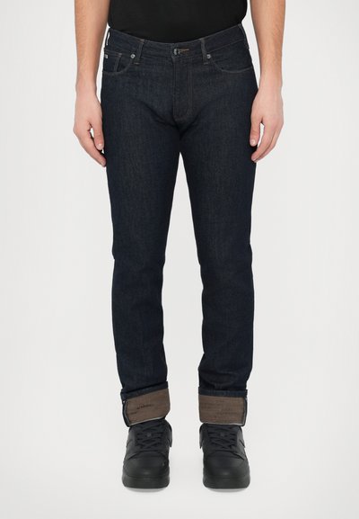 Dark blue denim jeans with a slim fit, featuring a brown cuff accent, contrast stitching, and a front button closure. Paired with black sneakers.