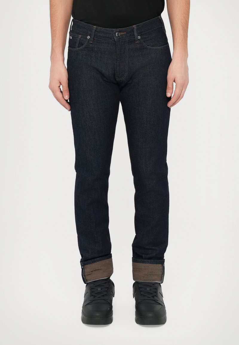 Sötétkék farmernadrág, karcsúsított szabással, barna szegéllyel, kontrasztos varrással és elöl gombos záródással. Fekete tornacipővel párosítva.