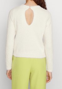 Pull blanc duveteux avec une fente en forme de goutte au dos et une fermeture par bouton, doté de manches longues et d'un ourlet droit. Porté avec un pantalon vert.