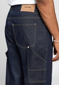 Pantalon cargo en denim foncé avec une étiquette en cuir beige, plusieurs poches, surpiqûres jaunes contrastantes et une coupe décontractée.