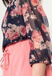 Blusa floral com tecido azul náutico transparente, apresentando grandes rosas pink e brancas, combinada com um fundo rosa texturizado e cordão.