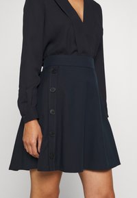 Marineblå kjol i mjukt tyg, med en åtsittande midja, utsvängd form och sex svarta knappar längs sidan. Slät textur och enkel design.