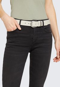 Vit skärp i läder med strukturerad yta, utrustad med spänne i silvermetall och flera hål för justering, bärs över svarta jeans.