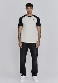 Camiseta de béisbol de manga corta en crema y negro con cuello redondo y logo, combinada con jeans negros y zapatillas blancas.
