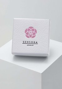 Balta tekstūrēta rotaslietu kaste ar rozā zieda logo un melnu tekstu, kas uzrakstīts "SENSORA JEWELRY", priekšpusē.