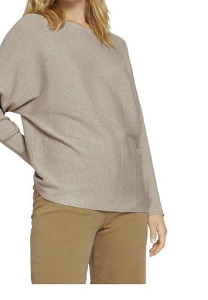 Mujer con un suéter holgado de punto beige con mangas largas y pantalones color beige claro, posando con una mano detrás de la espalda.