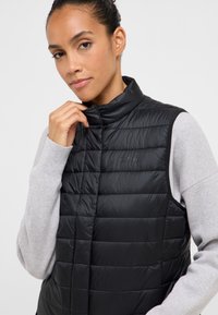 Czarna pikowana kamizelka puchowa z wysokim kołnierzem, przeszywanymi panelami i logo na piersi. Noszona na jasnoszarym swetrze.