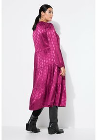 Fuchsia schichtkleid aus glänzendem Stoff, mit langen Ärmeln und einem hexagonalen Muster. Kombiniert mit knöchelhohen schwarzen Stiefeln mit Absätzen.