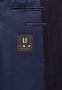 Jūras spēku auduma uzvalka iekšpuse, kas izceļas ar gludu satīna struktūru, divām kabatām un kvadrāta logo birku ar "BOGGI MILANO" baltajās burtiņās.